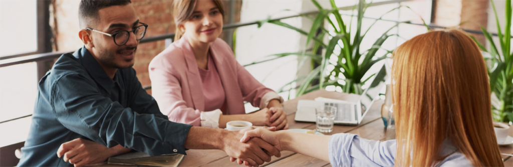 Two people shaking hands across a table in a meeting while a third person observes.