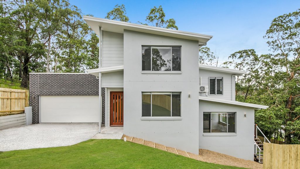 Modern two-story house with a garage, set on a sloped terrain with natural greenery in the background.