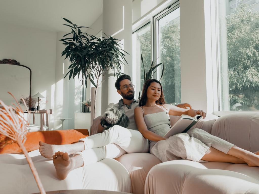 A couple relaxing on a couch with a book in a sunlit room with plants.