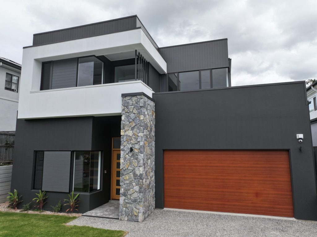 Modern two-story house with a flat roof, black and white exterior, stone column, large windows, wooden garage door, and a small front lawn.